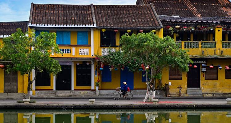 Cyclist wearing a conical hat passing colorful shutters and lanterns on a yellow riverside house in Hoi An.