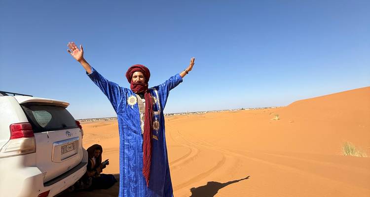 Berber guide in blue robes raising arms happily in the middle of Sahara sand dunes beside a 4x4 vehicle.