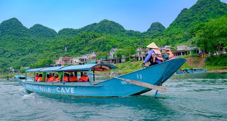 Blue tourist boat packed with lifejackets cruises past riverside village and lush karst hills in Phong Nha.