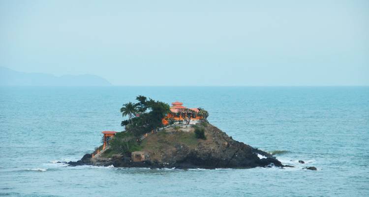 Tiny rocky islet topped by an orange temple and palm trees surrounded by turquoise sea.