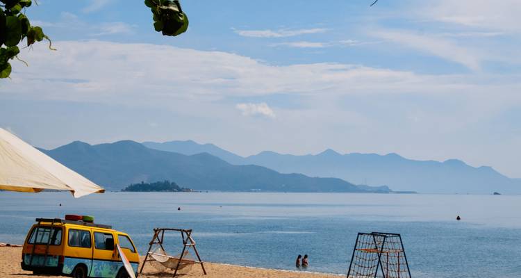 Plage de sable tranquille avec un van coloré, des balançoires et des montagnes brumeuses en arrière-plan de l'autre côté d'une eau calme.