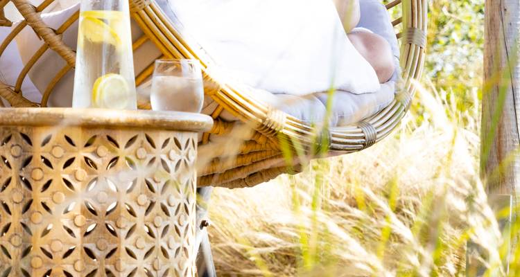 Gros plan lumineux d'une chaise suspendue en rotin avec des coussins à côté d'une table d'appoint à motifs tenant une carafe et un verre de citron pressé dans un jardin baigné de soleil.