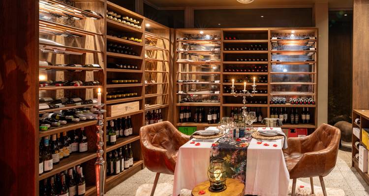 Cave à vin élégante avec des rayonnages en bois remplis de bouteilles encadrant une table dressée pour deux personnes, éclairée aux chandelles, avec des pétales de rose et une lanterne.