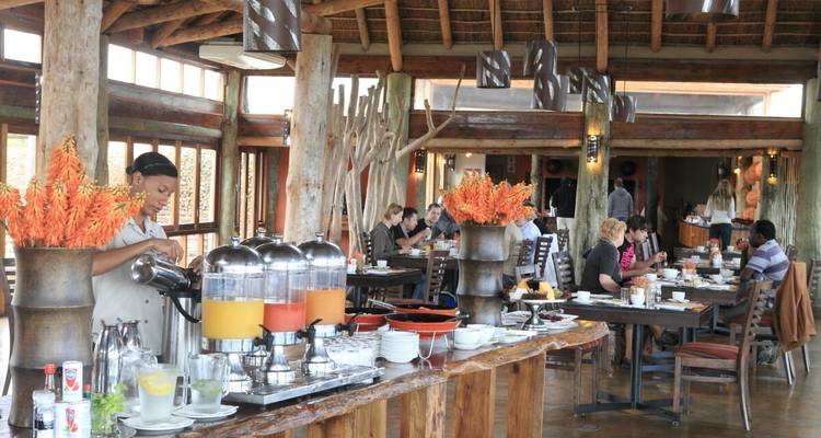 Restaurant de lodge rustique avec distributeurs de jus en libre-service et clients prenant leur petit-déjeuner sous un toit en chaume.