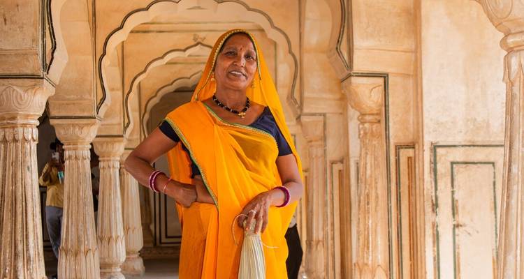 An older Indian woman in a golden sari stands proudly in a palace corridor framed by scalloped arches.