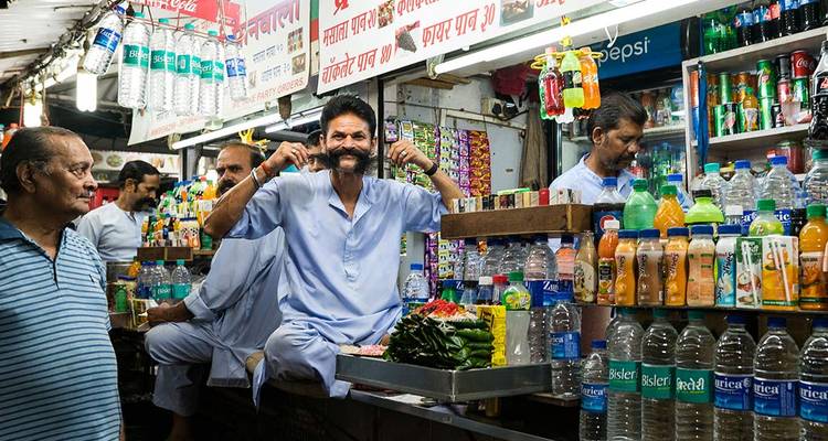 Lively market stall where a vendor playfully twirls his moustache amid rows of colorful bottled drinks.