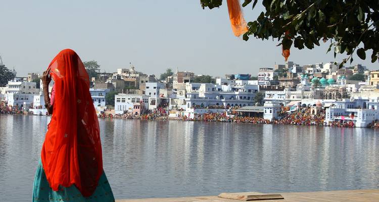 A woman in a bright red sari looks over the tranquil Pushkar Lake with white ghats and town in the background.