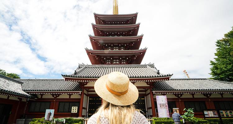 Bezoeker met stroohoed kijkt omhoog naar een vijfverdiepingse pagode tegen een felle hemel in Tokio.
