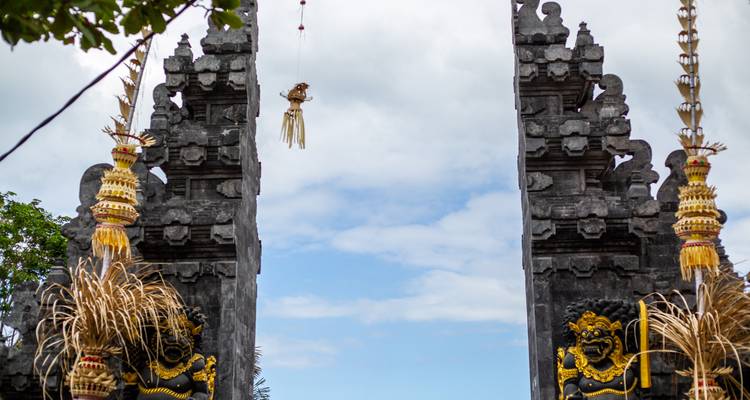 Puerta del templo balinés tradicional decorada con ofrendas que se extienden hacia un cielo parcialmente nublado.