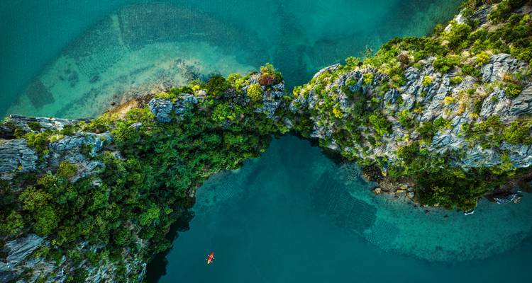 Vista aérea espectacular del mar esmeralda e islotes de piedra caliza dentada unidos por un puente de vegetación estrecho, con un pequeño kayak rojo abajo.