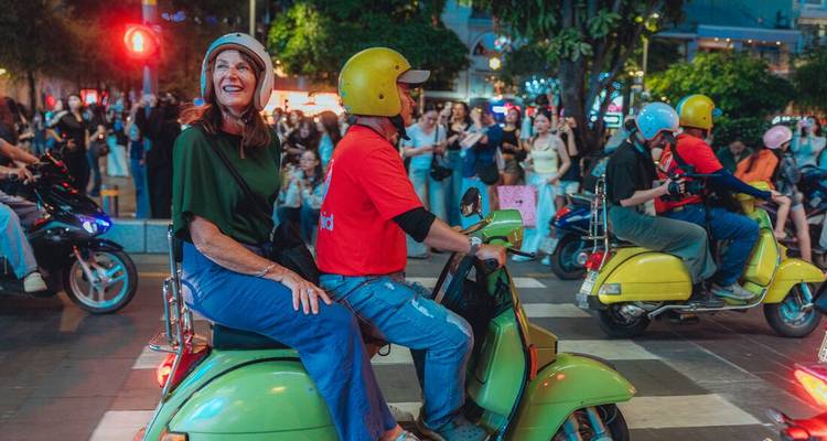 Female passenger smiles while riding pillion on a bright green scooter through a busy night intersection.