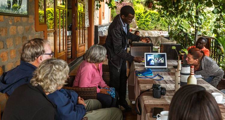 A guide presents wildlife photos to interested travellers gathered around a laptop on a lodge veranda.