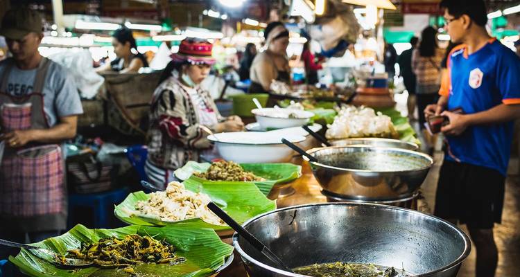 Étal de marché intérieur animé avec des plateaux de plats locaux servis sur des feuilles de bananier et des clients choisissant leur nourriture
