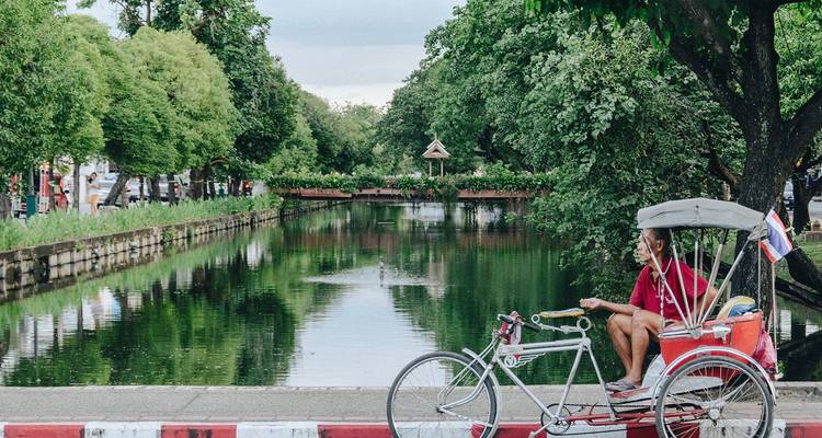 Le conducteur de pousse-pousse à pédales attend près d'un canal calme bordé d'arbres reflétant une verdure luxuriante
