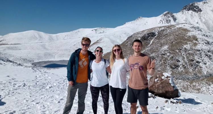 Four friends posing on a snowy volcanic slope with a blue crater lake and white peaks behind them.