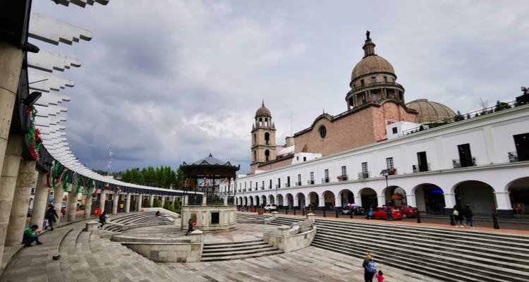 Wide plaza view of Toluca cathedral and colonnaded buildings with a dramatic cloudy sky.
