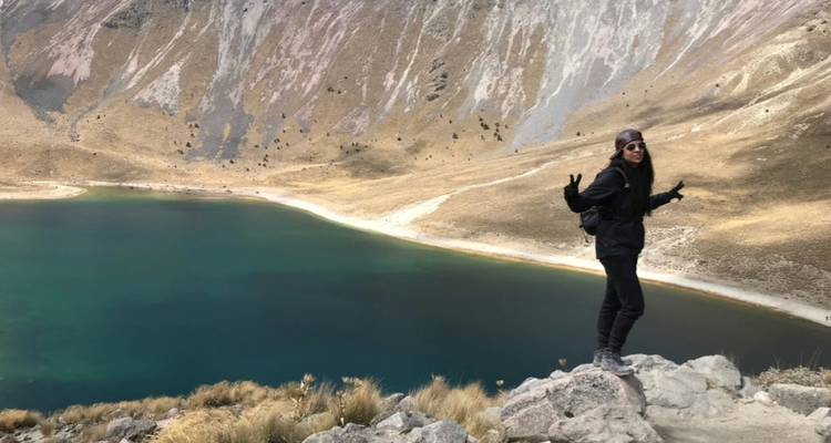 Solo hiker in dark clothing standing on rocky rim above a vivid crater lake in volcanic terrain.