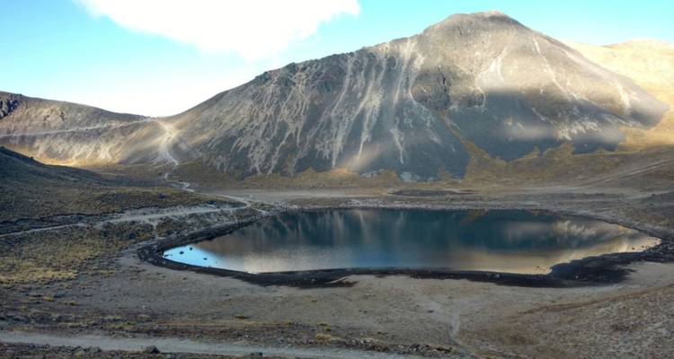 Still crater lake beneath rugged volcanic slopes under clear blue sky.