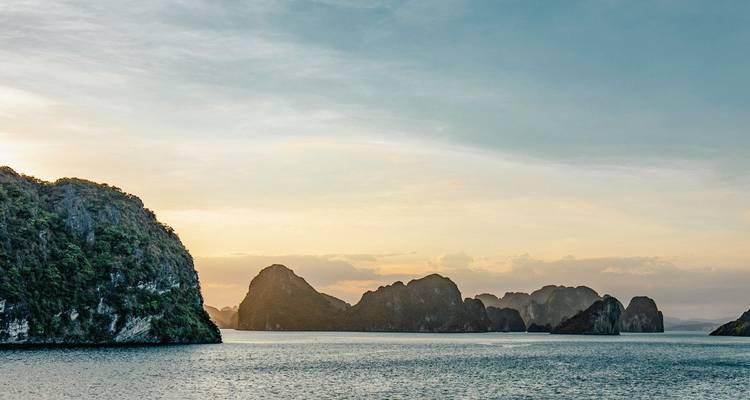 Karst limestone islands rise from the tranquil emerald waters of Halong Bay at pastel sunset.