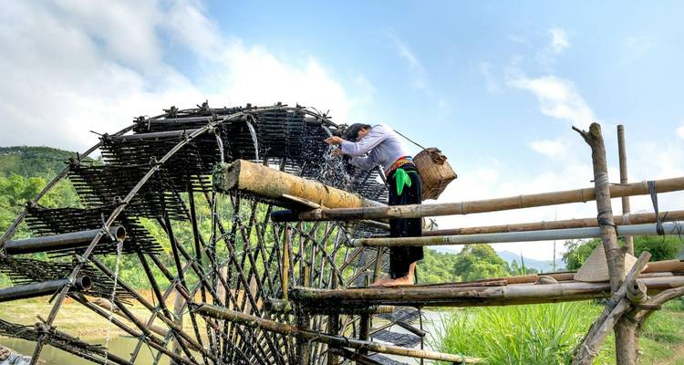 Person collecting water from a large bamboo water-wheel structure spanning a stream in lush countryside.