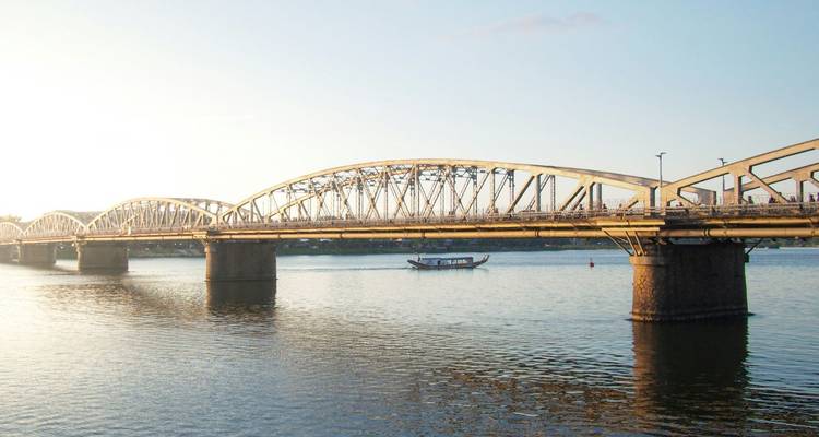 Lange metalen boogbrug die een brede rivier overspant in zacht avondlicht met een klein bootje dat eronder voorbijvaart