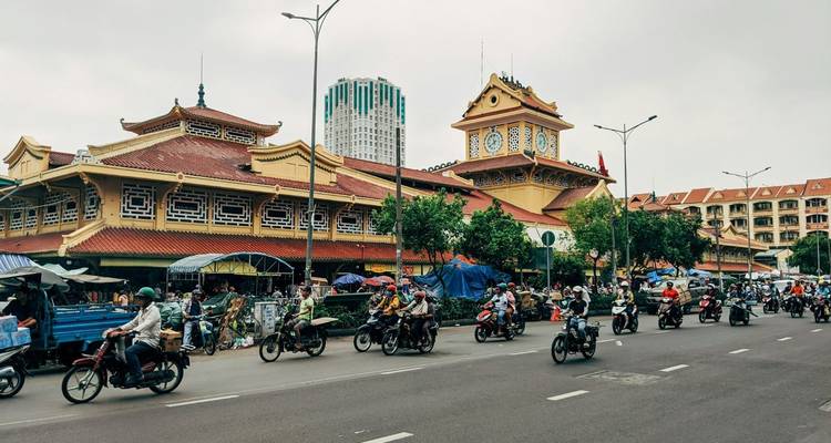 Levendig stedelijk marktgebouw met een klokkentoren omgeven door stromen brommers- en motorfietsverkeer op een stadsweg