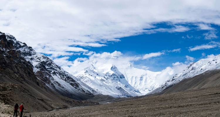 Two trekkers approach a dramatic snow-covered Himalayan range beneath a vivid blue sky with scattered clouds.