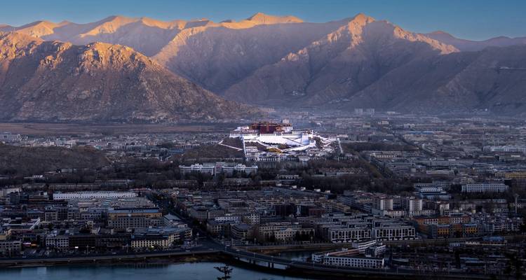 Wide aerial of Lhasa city at dawn with Potala Palace centered below glowing mountain peaks.