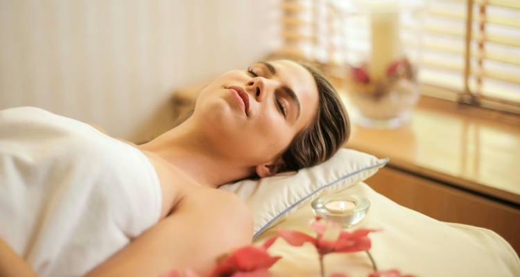 Young woman wrapped in a towel relaxes on a spa bed with candle and flowers nearby.
