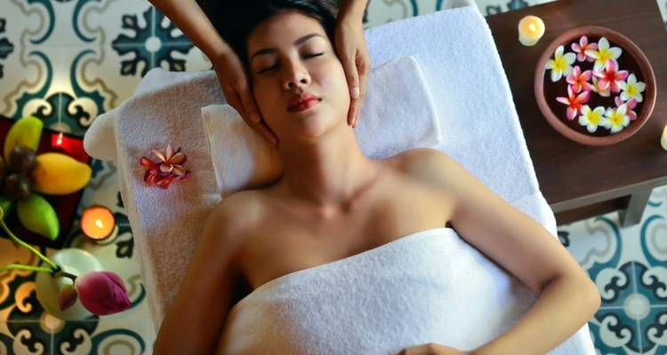 Spa therapist gently massages a woman’s temples as she rests on a towel surrounded by candles and flowers.