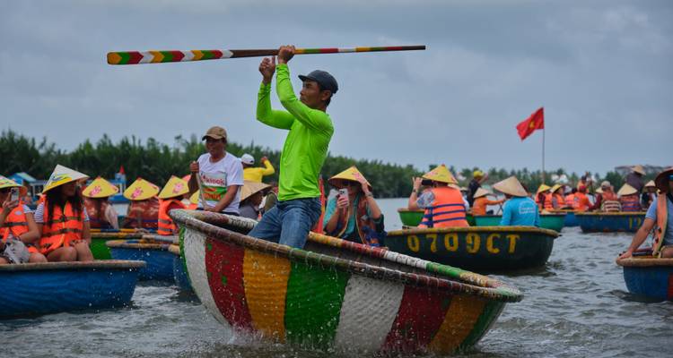 Man energetically paddles a colourful round basket boat while crowds of tourists watch from their own boats.