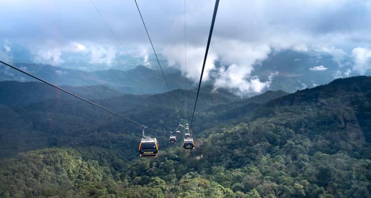 Long cable-car line descends over green forested mountains into cloud gaps.