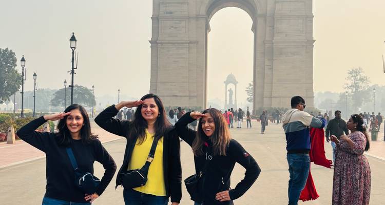 Three women saluting playfully in front of India Gate with crowds in the background