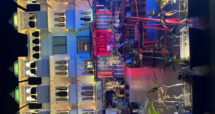A palace-style hotel courtyard glows with colourful lights, potted plants and alfresco seating against a night sky.