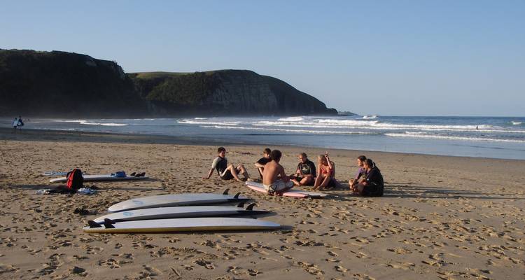 Small group of surfers seated on the sand beside their boards facing rolling waves.