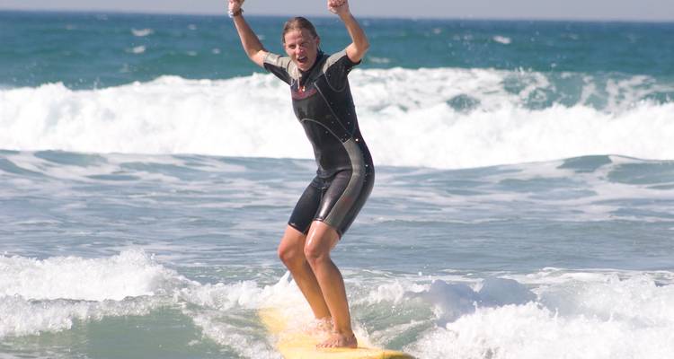 Smiling surfer raises arms in triumph while riding a white-water wave on a sunny day.