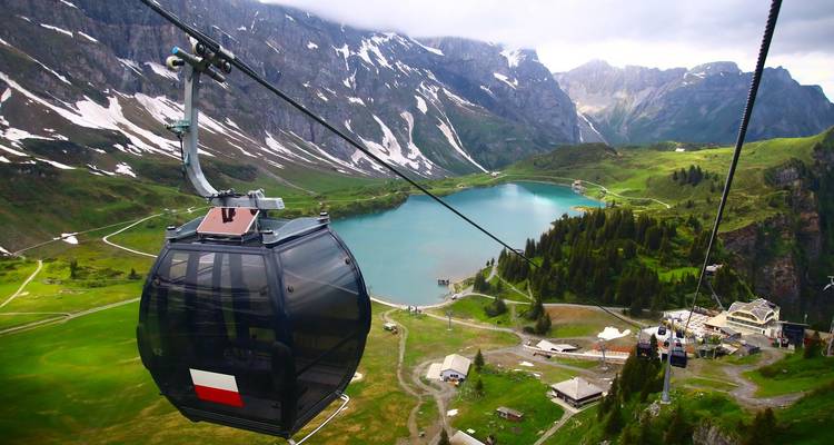 Teleférico deslizándose sobre un lago alpino esmeralda rodeado de montañas con parches de nieve.