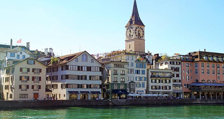 Fila de edificios en tonos pastel a orillas del río y una iglesia con campanario reflejadas en aguas verdes en un día soleado.