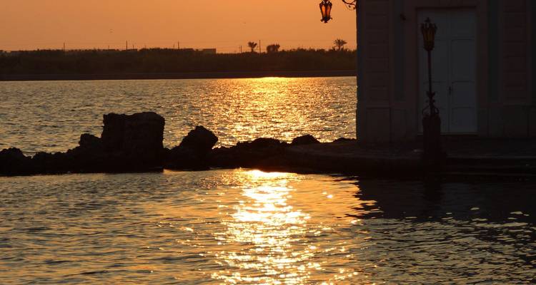 Goldener Sonnenuntergang, der sich auf ruhigem Wasser neben einem silhouettierten Gebäude und Laternen spiegelt.