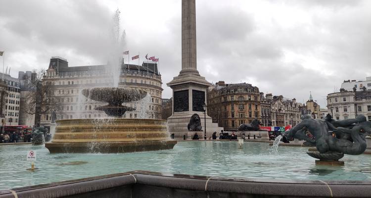 Close view of Trafalgar Square fountain with National Gallery façade and cloudy skies.