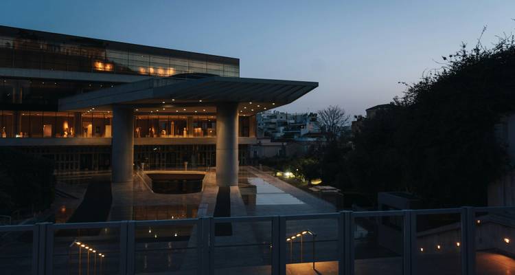 Evening exterior of the modern Acropolis Museum with lights reflecting on sleek wet pavement.