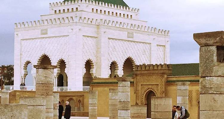 The white marble Mausoleum of Mohammed V stands amid ancient stone columns with a few visitors walking nearby.