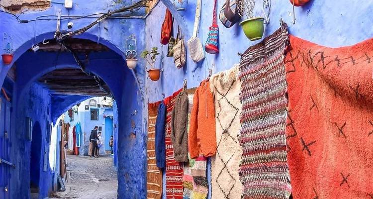 Vibrant hand-woven rugs hang along narrow cobalt-blue alleyways in Chefchaouen as two tourists explore in the distance.