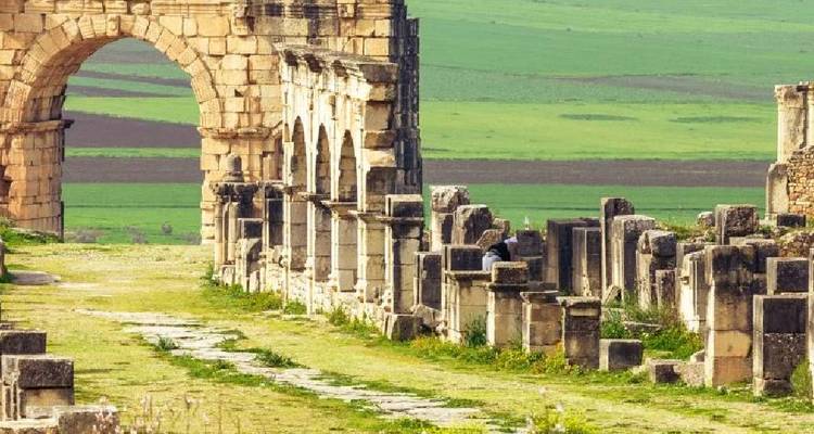 Stone arches and crumbling columns of the ancient Roman city of Volubilis stand amid rolling green fields.