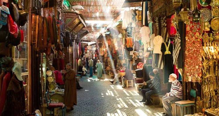 A lively, sun-dappled souk passage is lined with colourful textiles and brassware as locals and visitors browse.