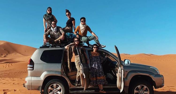 A group of friends poses atop a 4x4 vehicle parked on orange desert sand under a clear blue sky.