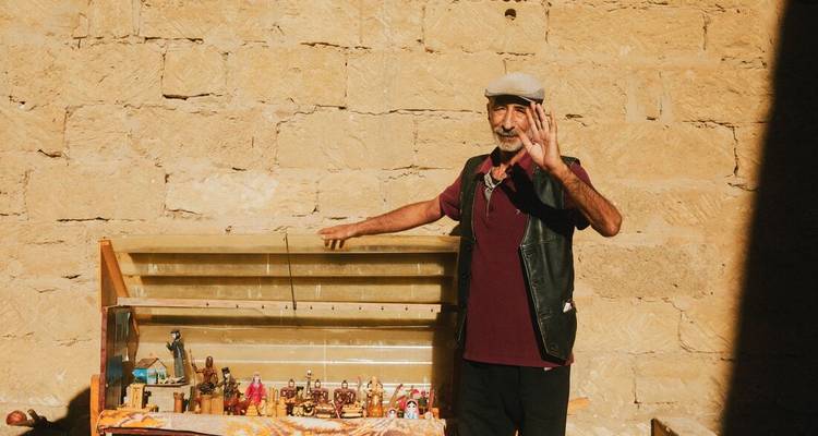 An older man in casual clothes waves while standing beside a small outdoor souvenir stall against a sun-lit stone wall.