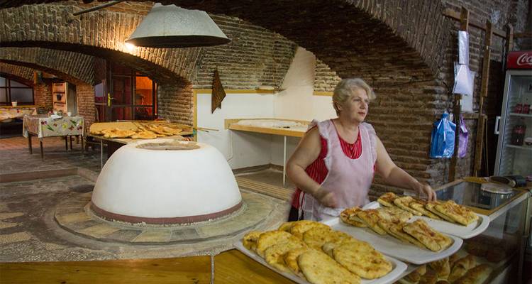 An older woman prepares traditional flatbread beside a clay oven inside a rustic brick bakery.