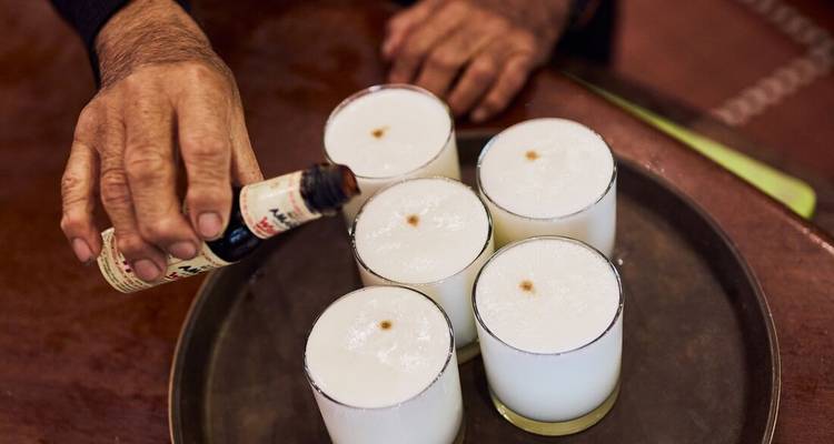 Close-up of a hand adding bitters to a tray of frothy pisco sour cocktails.