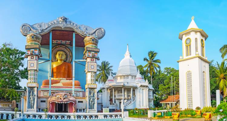 Brightly painted Buddhist temple complex with giant seated Buddha and white stupa under clear sky.